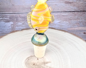 May include: A glass bottle stopper featuring a stylised fish ornament with yellow and white swirling patterns. The fish has a small dark eye and translucent fins. The stopper base consists of a smooth, rounded teal glass section attached to a tapered cork.