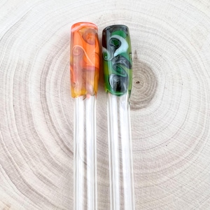 Puede incluir: Dos pajitas de vidrio con puntas decorativas. Una pajita tiene un diseño de remolino naranja y amarillo, y la otra tiene un diseño de remolino verde, negro y blanco. Las pajitas son transparentes y están sobre una superficie de madera.
