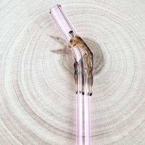 Può includere: Una cannuccia di vetro rosa con un design decorativo a mano marrone. La cannuccia è angolata e presenta una superficie chiara e liscia. Il design a mano aggiunge un tocco unico all'aspetto della cannuccia.