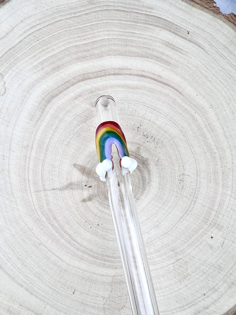 May include: A clear glass drinking straw featuring a decorative rainbow arch fused to the exterior. The rainbow is composed of red, orange, yellow, green, blue, and purple bands, with small opaque white glass clouds attached at each base end.