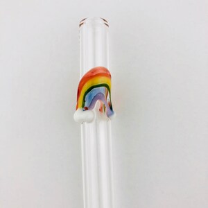 May include: A clear glass drinking straw featuring a decorative three-dimensional rainbow accent fused to the side. The rainbow is composed of red, orange, yellow, green, blue, and purple glass stripes with small white glass clouds at each base.
