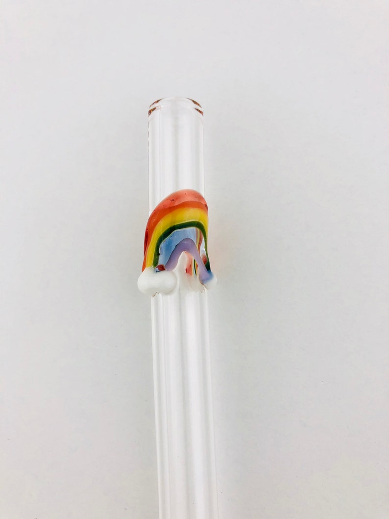 May include: A clear glass drinking straw featuring a decorative three-dimensional rainbow accent fused to the side. The rainbow is composed of red, orange, yellow, green, blue, and purple glass stripes with small white glass clouds at each base.