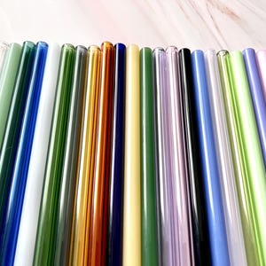 May include: A collection of colorful glass straws in various shades, including green, blue, gold, and purple. The straws are arranged in a row, showcasing their different colors and transparency. These reusable straws are a sustainable alternative to plastic.