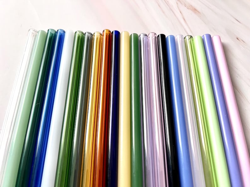 May include: A collection of colorful glass straws in various shades, including green, blue, gold, and purple. The straws are arranged in a row, showcasing their different colors and transparency. These reusable straws are a sustainable alternative to plastic.