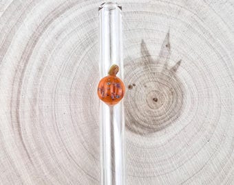 Può includere: Una cannuccia in vetro trasparente con un piccolo decoro in vetro a forma di zucca arancione applicato sul lato. La zucca presenta sottili macchie marroni e bianche e un piccolo picciolo verde sulla parte superiore, integrato nella cannuccia.