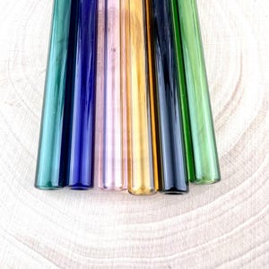 May include: An assortment of glass straws in a range of colours, including green, blue, pink, and gold. The straws are displayed side by side, emphasizing their different shades and cylindrical form. The image is shot from above, highlighting the smooth, transparent texture of the glass.