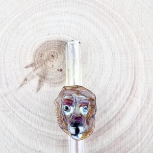 May include: A clear glass pipe featuring a sculpted lion head ornament. The lion is crafted from brown, tan, and white glass with distinct facial features, including dark eyes and a white muzzle, attached to the cylindrical stem of the pipe.