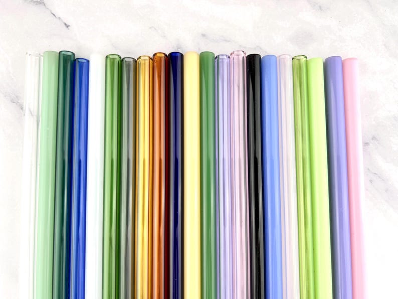 May include: A collection of colorful glass straws in various shades, including green, blue, yellow, and pink. The straws are arranged vertically, showcasing their smooth, cylindrical design. The image is taken against a white marble background.
