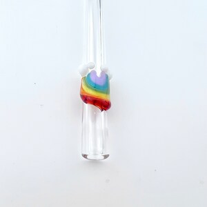 May include: A clear glass straw featuring a decorative accent shaped like a rainbow. The rainbow design consists of layered bands of purple, blue, green, yellow, orange, and red glass, topped with small, opaque white glass spheres on each side.