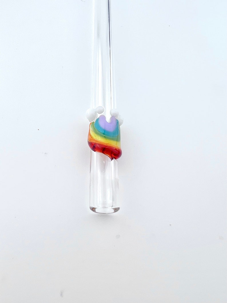 May include: A clear glass straw featuring a decorative accent shaped like a rainbow. The rainbow design consists of layered bands of purple, blue, green, yellow, orange, and red glass, topped with small, opaque white glass spheres on each side.