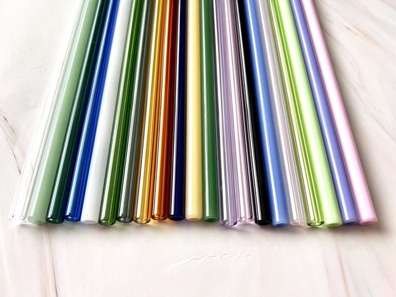 May include: A collection of colorful glass straws in various shades, including green, blue, yellow, and pink. The straws are arranged in a diagonal pattern, showcasing their vibrant colors and smooth, cylindrical shape. These reusable straws are a sustainable alternative.
