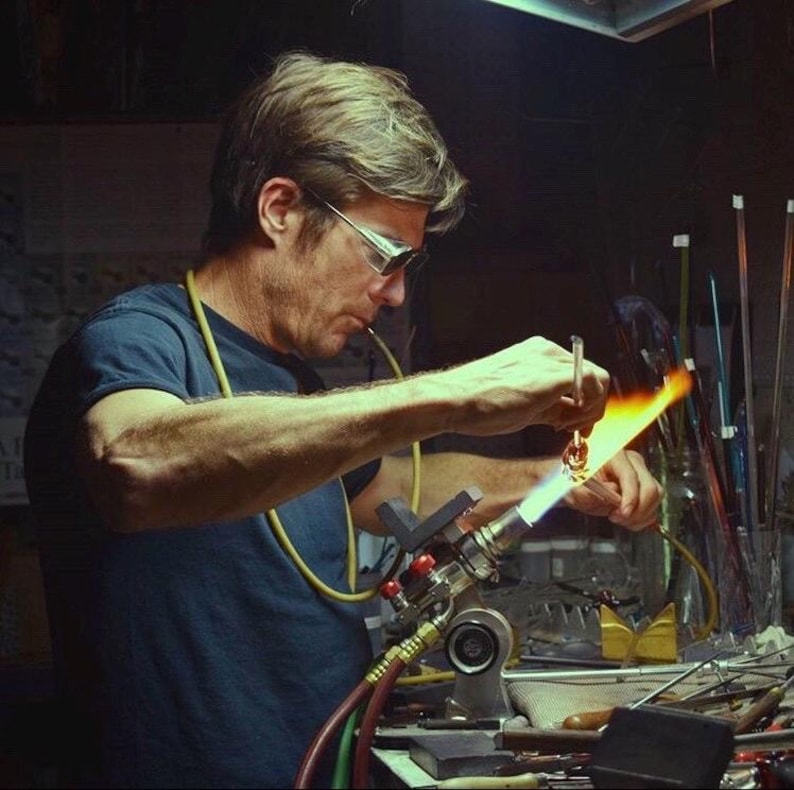 May include: A person working with a torch, creating a glass art piece. The person is wearing safety glasses and a dark blue t-shirt. The flame is bright yellow and orange, illuminating the workspace.
