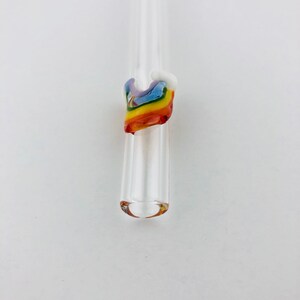 May include: A clear glass drinking straw featuring a decorative accent near the top. The accent is shaped like a small, multi-colored rainbow made of fused glass, displaying bands of red, orange, yellow, green, blue, and violet colors.