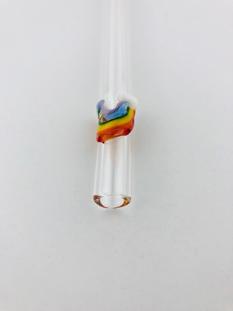 May include: A clear glass drinking straw featuring a decorative accent near the top. The accent is shaped like a small, multi-colored rainbow made of fused glass, displaying bands of red, orange, yellow, green, blue, and violet colors.