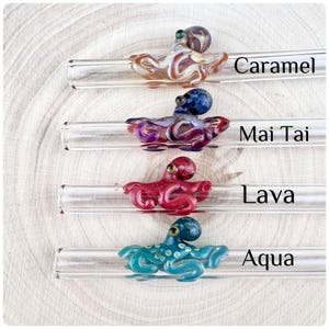 May include: Four clear glass drinking straws, each featuring a decorative glass octopus figurine. The octopuses are coloured caramel, mai tai, lava, and aqua, with corresponding text labels printed next to each straw on the right side.