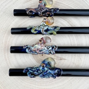 May include: Four black glass pipes, each decorated with a colourful, detailed glass octopus. The octopuses feature various colours, including blue, purple, green, and yellow, with intricate detailing. The pipes rest on a light wood surface.