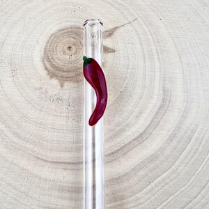 May include: A clear glass straw with a red chili pepper design. The chili pepper is a vibrant red with a green stem. The straw is resting on a light brown wooden surface.