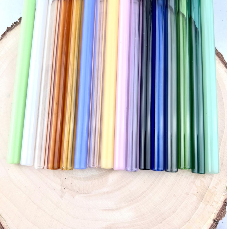 May include: A collection of colorful glass straws in various shades, including green, clear, brown, blue, yellow, and purple. The straws are arranged vertically on a wooden surface, showcasing their different hues and transparency. These reusable straws are a sustainable alternative.