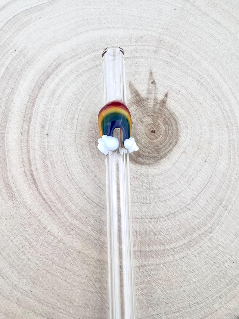 May include: A clear glass drinking straw featuring a decorative, fused glass rainbow arch design. The rainbow is composed of red, orange, yellow, green, blue, and violet bands, with small, opaque white glass clouds attached at each end of the arch.