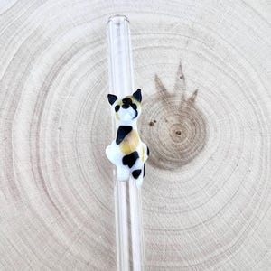May include: A clear glass drinking straw featuring a small, three-dimensional calico cat figure attached to the side. The cat is crafted from white, black, and tan glass, showing a seated posture with pointed ears and distinct color patches.