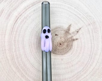 Può includere: Una cannuccia dritta in vetro grigio scuro traslucido con una piccola figura di fantasma tridimensionale di colore viola chiaro applicata all'esterno. Il fantasma ha due occhi ovali neri e una piccola bocca circolare nera.
