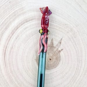 May include: A glass stirring rod featuring a sculpted squid topper. The squid is rendered in shades of deep red and pink with prominent yellow eyes and textured tentacles. The rod itself is a translucent dark green, extending from the squid base.