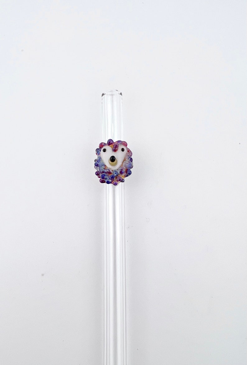 Peut inclure: Une paille &agrave; boire en verre transparent avec une perle en verre violette et blanche en haut. La perle est en forme de c&oelig;ur et a un visage blanc avec des yeux noirs et un nez dor&eacute;.