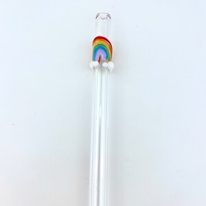 May include: A clear glass drinking straw featuring a small, decorative glass rainbow accent near the top. The rainbow is composed of red, orange, yellow, green, blue, and purple bands, with two small white glass clouds attached at the base.
