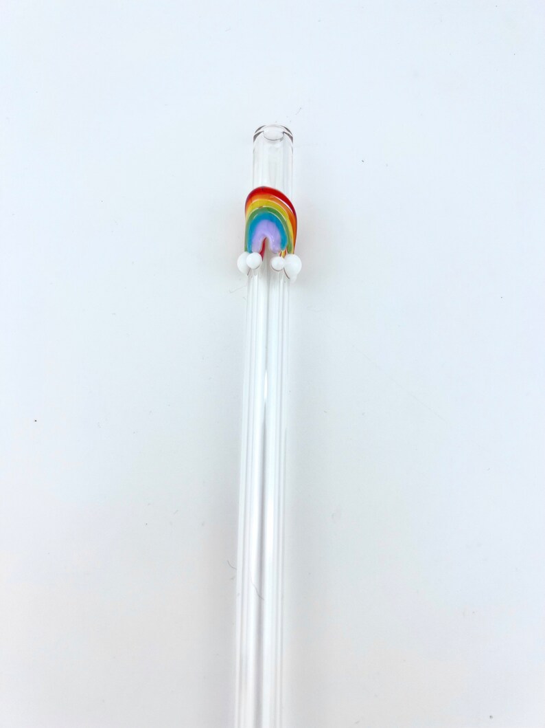 May include: A clear glass drinking straw featuring a small, decorative glass rainbow accent near the top. The rainbow is composed of red, orange, yellow, green, blue, and purple bands, with two small white glass clouds attached at the base.