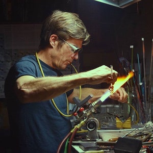 Peut inclure: Un souffleur de verre utilise un chalumeau pour chauffer une tige de verre, cr&eacute;ant une flamme orange vif. Le souffleur de verre porte des lunettes de s&eacute;curit&eacute; et un t-shirt bleu. Le souffleur de verre travaille &agrave; un &eacute;tabli avec divers outils et mat&eacute;riaux.