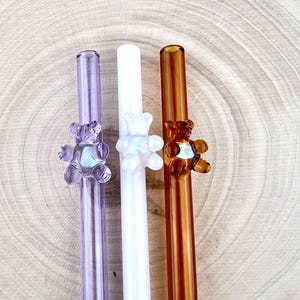 May include: Three glass straws with teddy bear accents. One straw is purple, one is white, and the third is amber. The straws are displayed on a light brown, wood-grain surface.