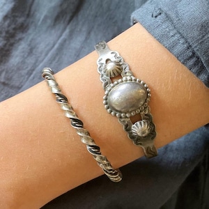 May include: Two silver-toned metal bracelets. One is a simple twisted rope design. The other is a wider cuff featuring a central oval cabochon stone surrounded by a beaded border, flanked by two smaller circular embossed accents on a textured band.