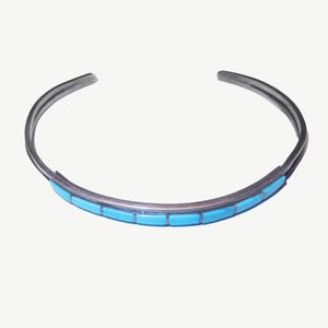 May include: A silver-toned cuff bracelet featuring a series of rectangular, bright turquoise-colored inlays arranged in a single row along the front center of the band. The metal band has a smooth, polished finish and an open-ended design.