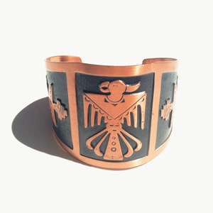 Pode incluir: Uma pulseira de cobre com um design preto e cobre de um pássaro estilizado. O pássaro está no centro da pulseira e é rodeado por uma borda de cobre.