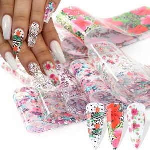 Puede incluir: Un conjunto de l&aacute;minas de transferencia de nail art floral. Las l&aacute;minas son transparentes con una variedad de patrones florales en rosa, rojo y blanco. Las l&aacute;minas se muestran sobre un fondo blanco con un maniqu&iacute; de mano que lleva nail art.