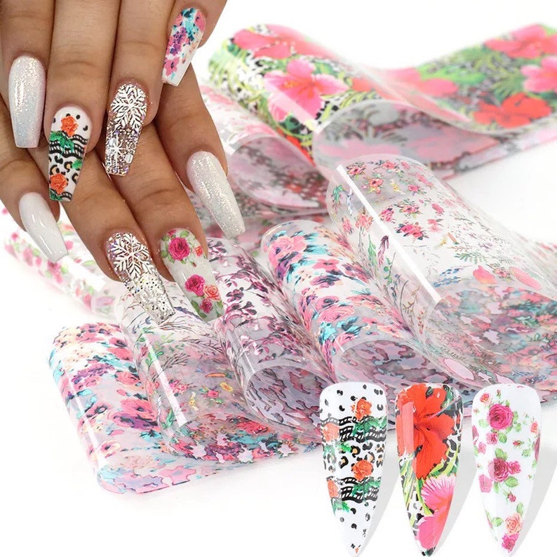Puede incluir: Un conjunto de l&aacute;minas de transferencia de nail art floral. Las l&aacute;minas son transparentes con una variedad de patrones florales en rosa, rojo y blanco. Las l&aacute;minas se muestran sobre un fondo blanco con un maniqu&iacute; de mano que lleva nail art.