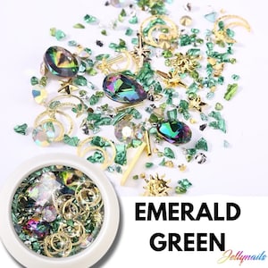 May include: A round container filled with green, gold, and iridescent nail art embellishments. The container is white with a clear lid. The embellishments include small green and gold glitter, iridescent gems, and gold crescent moons. The text "EMERALD GREEN" is written in large white letters below the container.