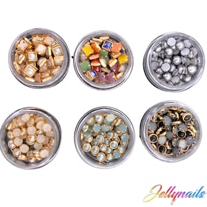 May include: Six small jars filled with various colorful and metallic nail embellishments. The jars are arranged in two rows of three. The embellishments include small squares, round pearls, and round rhinestones.