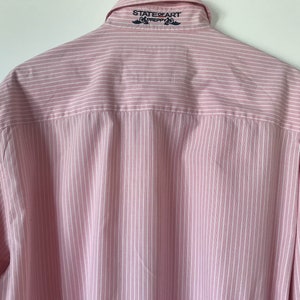 May include: A pink and white striped long-sleeve button-down shirt with the text "STATE OF ART PREPPY" embroidered on the back collar.