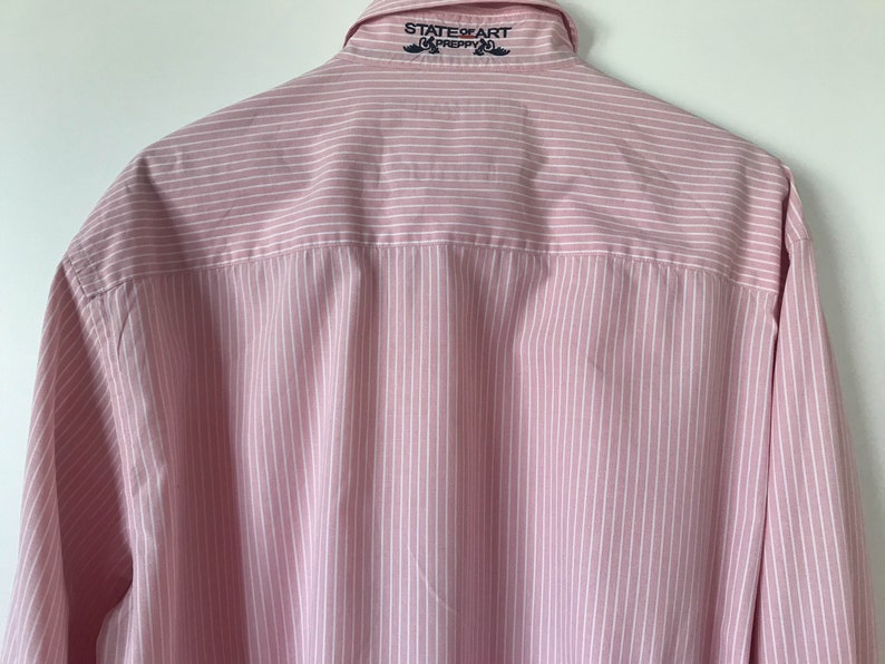 May include: A pink and white striped long-sleeve button-down shirt with the text "STATE OF ART PREPPY" embroidered on the back collar.