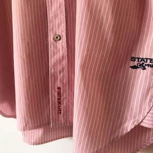 May include: Pink and white striped button-down shirt with the text "State of Art" embroidered on the left side and "State of Art Preppy" embroidered on the right side.