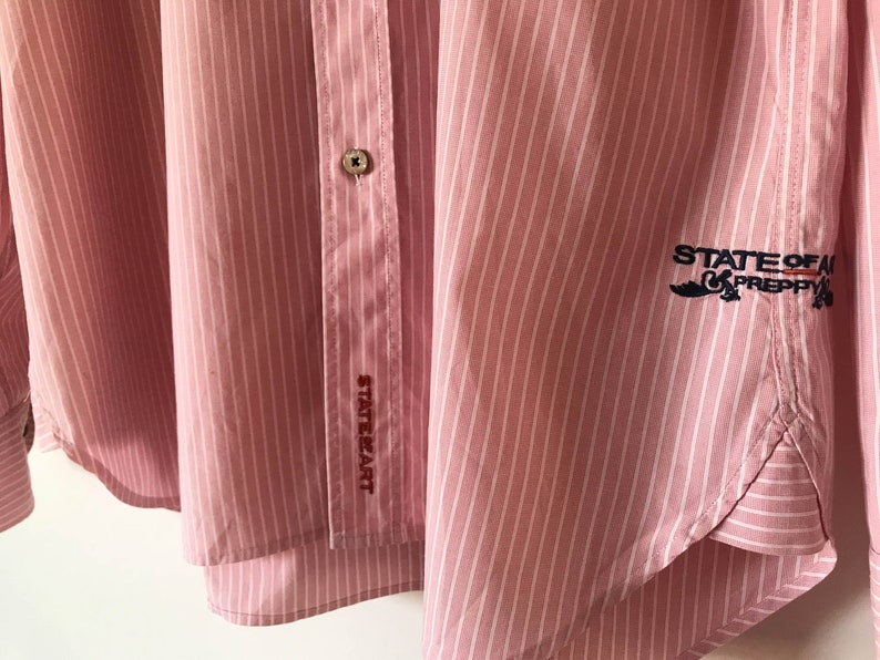 May include: Pink and white striped button-down shirt with the text "State of Art" embroidered on the left side and "State of Art Preppy" embroidered on the right side.