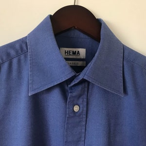 May include: A blue long-sleeved button-down shirt with a collar. The shirt has a label that reads "HEMA CLASSIC".