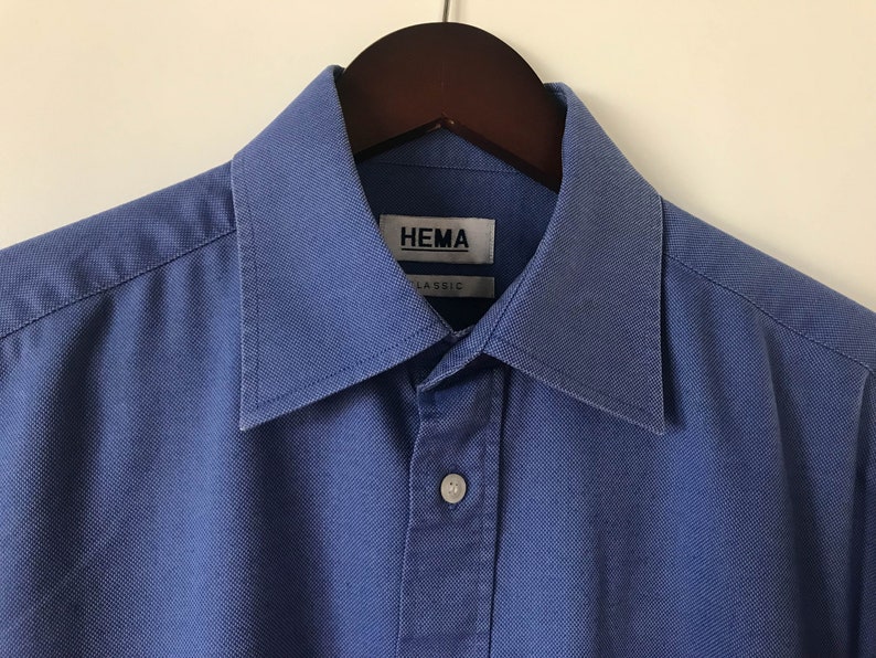 May include: A blue long-sleeved button-down shirt with a collar. The shirt has a label that reads "HEMA CLASSIC".