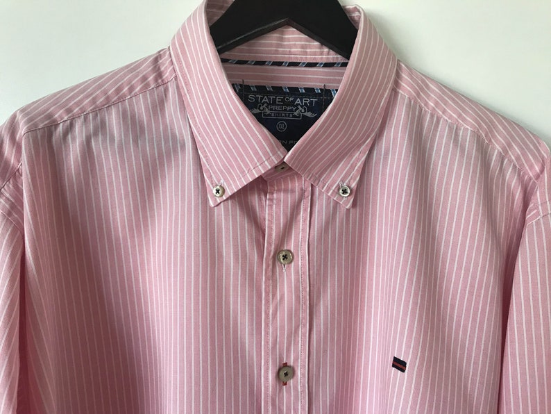 May include: A pink and white striped long-sleeve button-down shirt with a State of Art Preppy Shirts label. The shirt has a button-down collar and a small logo on the left chest.