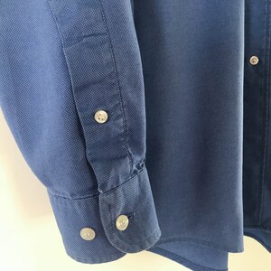 May include: A blue long-sleeved button-down shirt with a textured fabric. The shirt has a button-up collar and a single button on the right side of the chest.