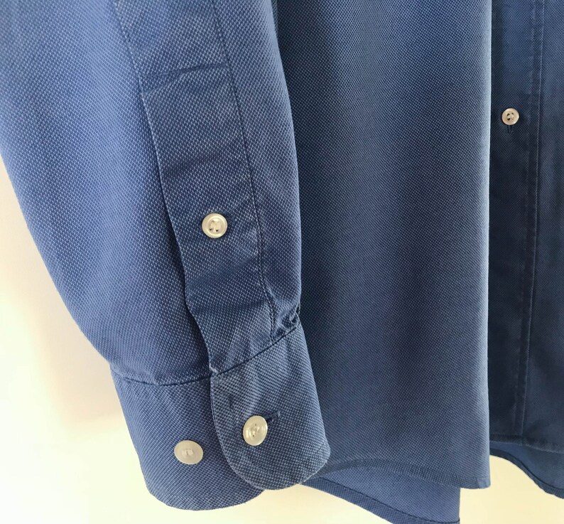 May include: A blue long-sleeved button-down shirt with a textured fabric. The shirt has a button-up collar and a single button on the right side of the chest.