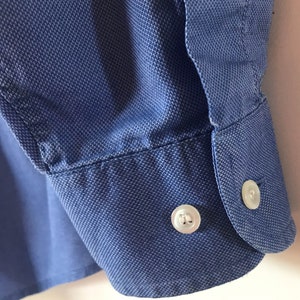 May include: A blue long-sleeved shirt with a button cuff. The button is white with a raised design.
