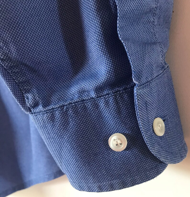 May include: A blue long-sleeved shirt with a button cuff. The button is white with a raised design.