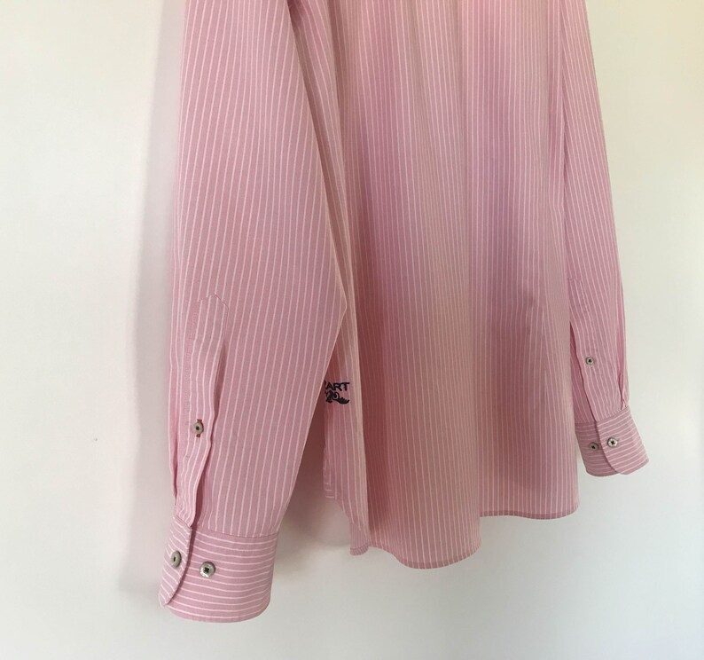 May include: A pink and white striped long-sleeved shirt with a small embroidered logo on the left sleeve. The logo says "HART OF" with a small mustache.